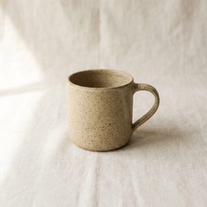 A sand-coloured speckled stoneware mug on a linen cloth